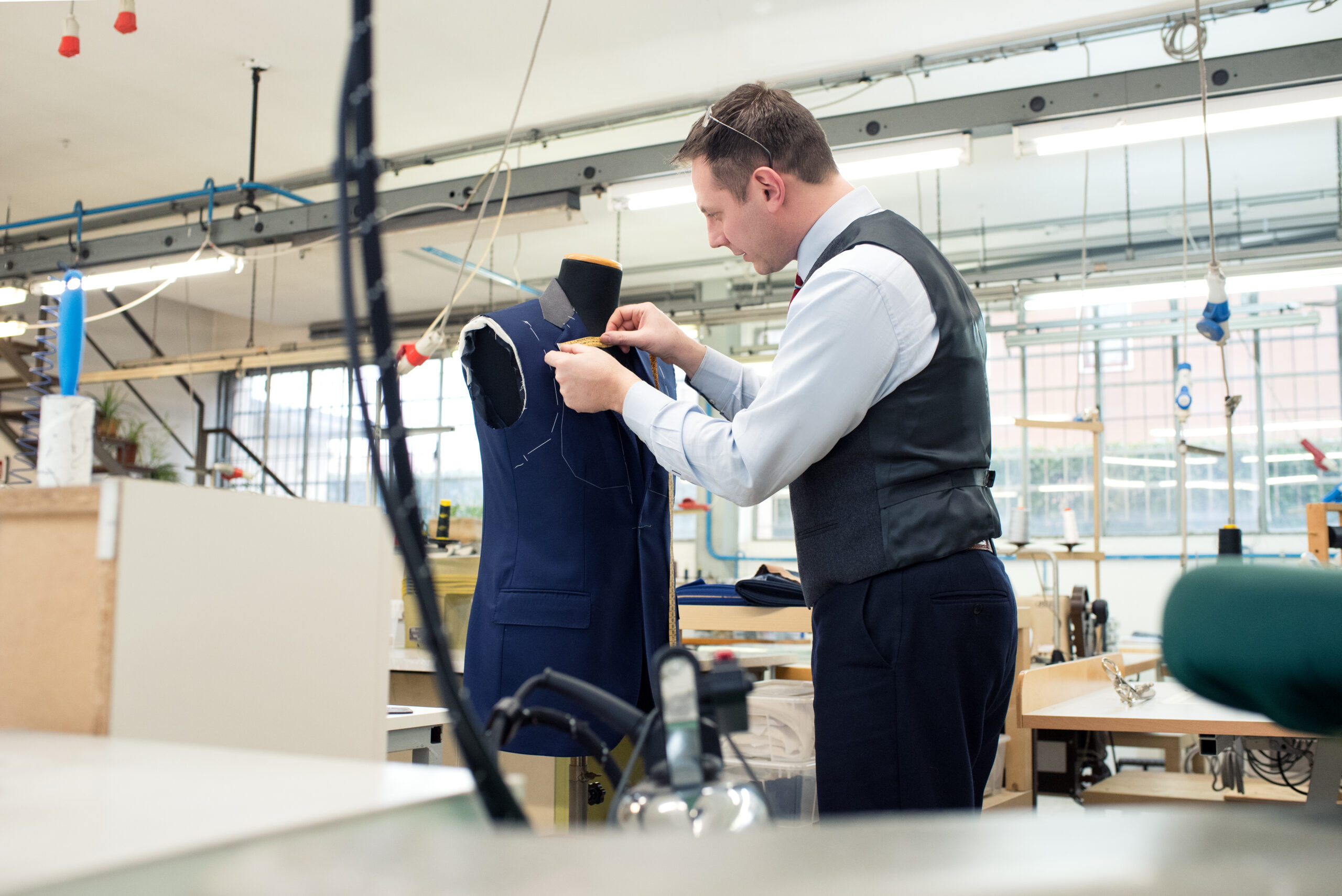 Tailor standing working on a blue jacket hanging on a mannequin in a bright spacious modern workshop viewed across a table