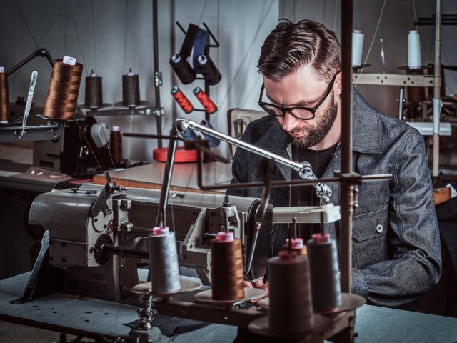 Attractive man seamstress is sewing clothes by vintage sewing machine. He is wearing glasses and denim. There are a lot of spools of threads at backgroud.
