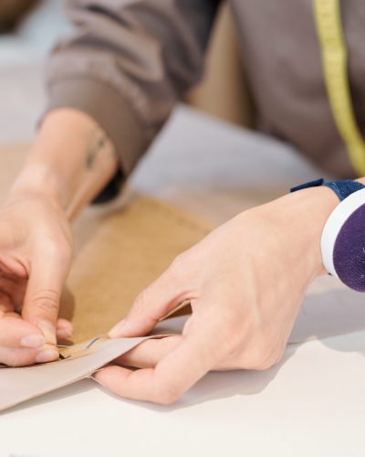 Hands of young seamstress pinning cut sketch to textile while working over new model of clothes in workshop