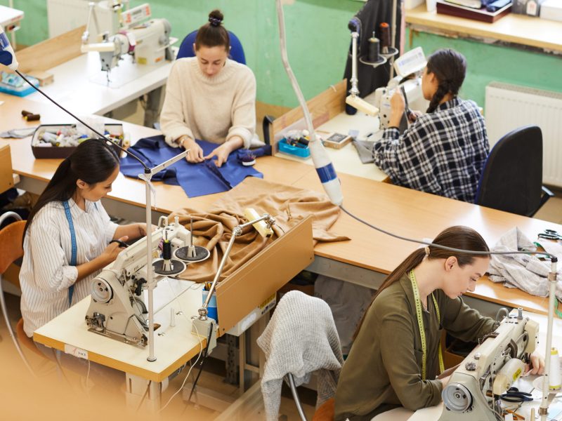 Group of young women sitting at the table and sewing clothes using their sewing machines in atelier