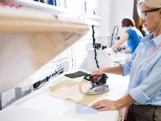 Portrait of mature woman ironing clothes with old fashioned metal iron in traditional atelier studio, copy space