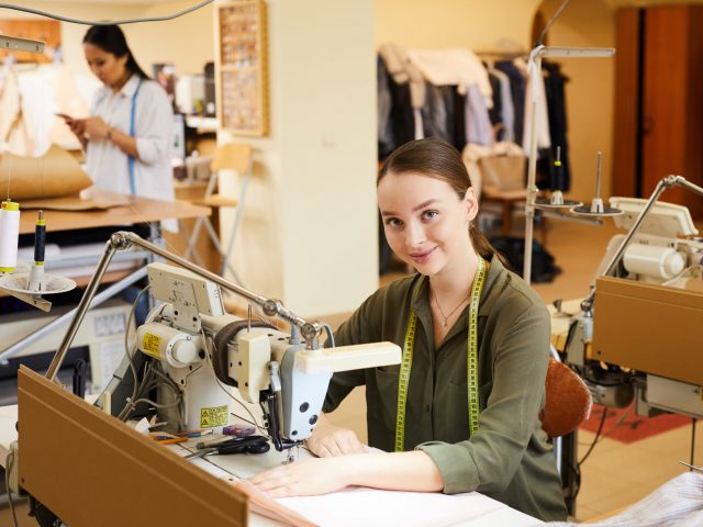 Portrait of young woman sitting at the table and using sewing machine in her work in atelier