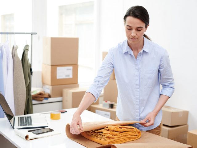Young manager of online shop wrapping folded yellow trousers into paper while packing it before sending to client
